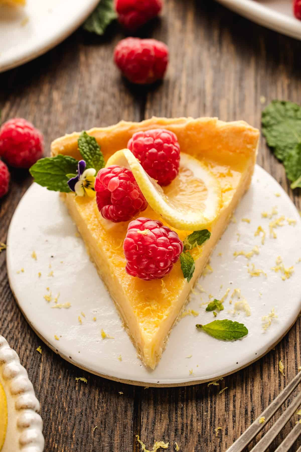 Lemon Tart A slice of lemon tart topped with fresh raspberries, a lemon slice, mint leaves, and a small edible flower.