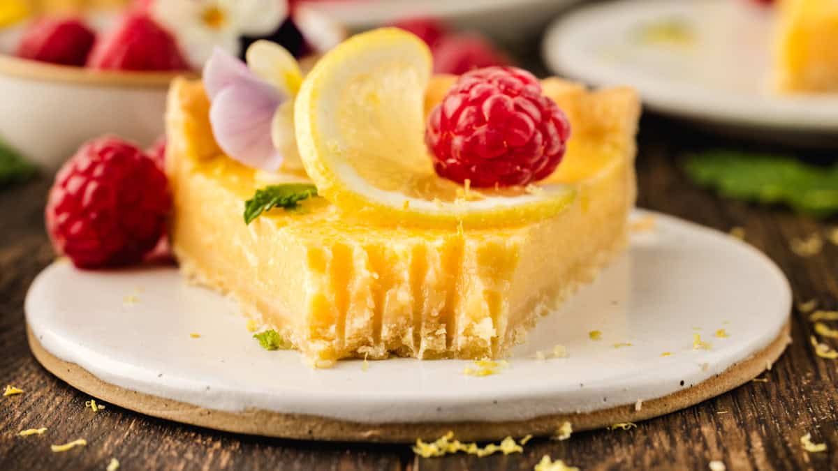 Lemon Tart A slice of lemon tart garnished with a lemon slice, raspberry, edible flowers, and mint sits on a white plate.
