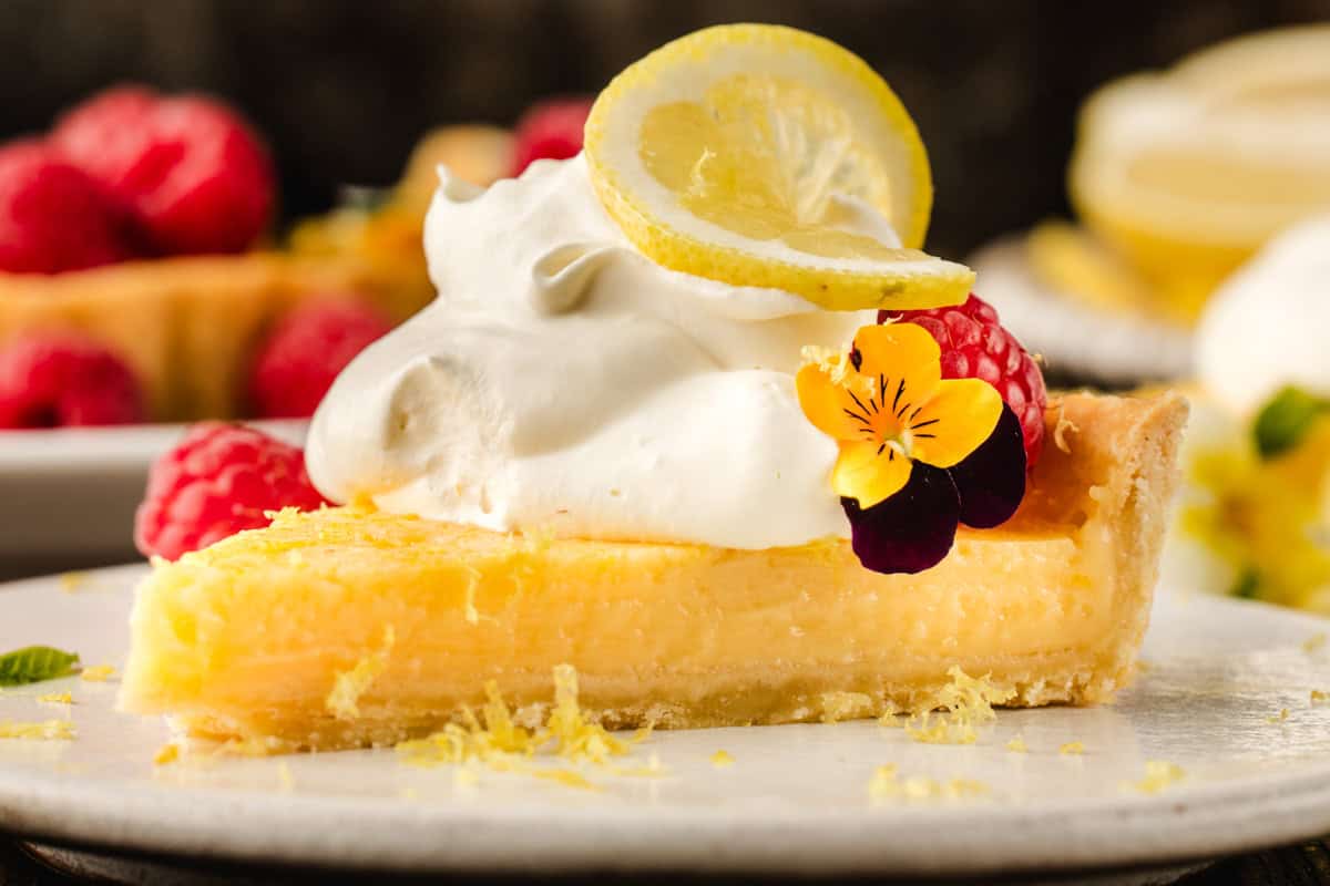 Lemon Tart A slice of lemon tart topped with whipped cream, a lemon slice, raspberries, and edible flowers sits on a white plate.