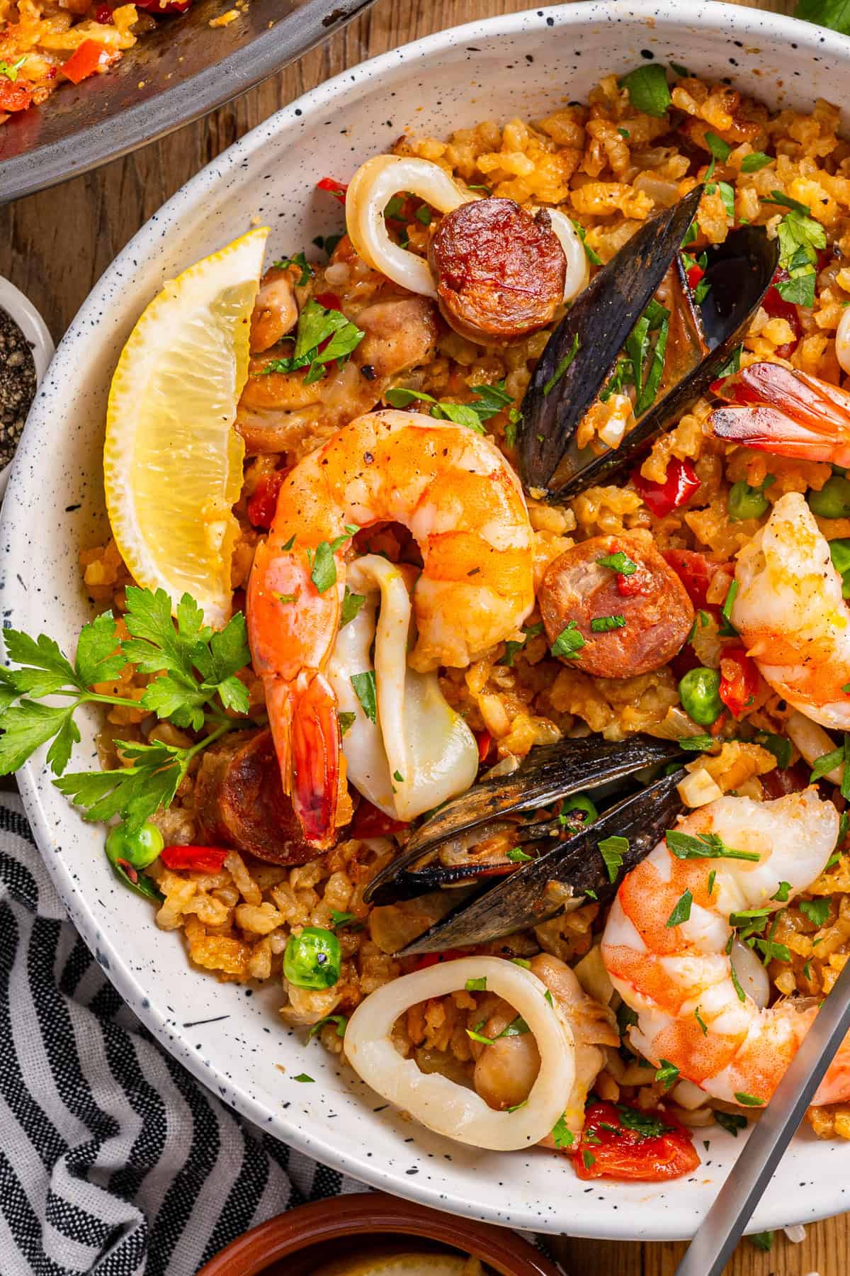 Paella A close-up of a bowl of seafood paella with shrimp, mussels, calamari, sausage, peas, red peppers, rice, and a lemon wedge.