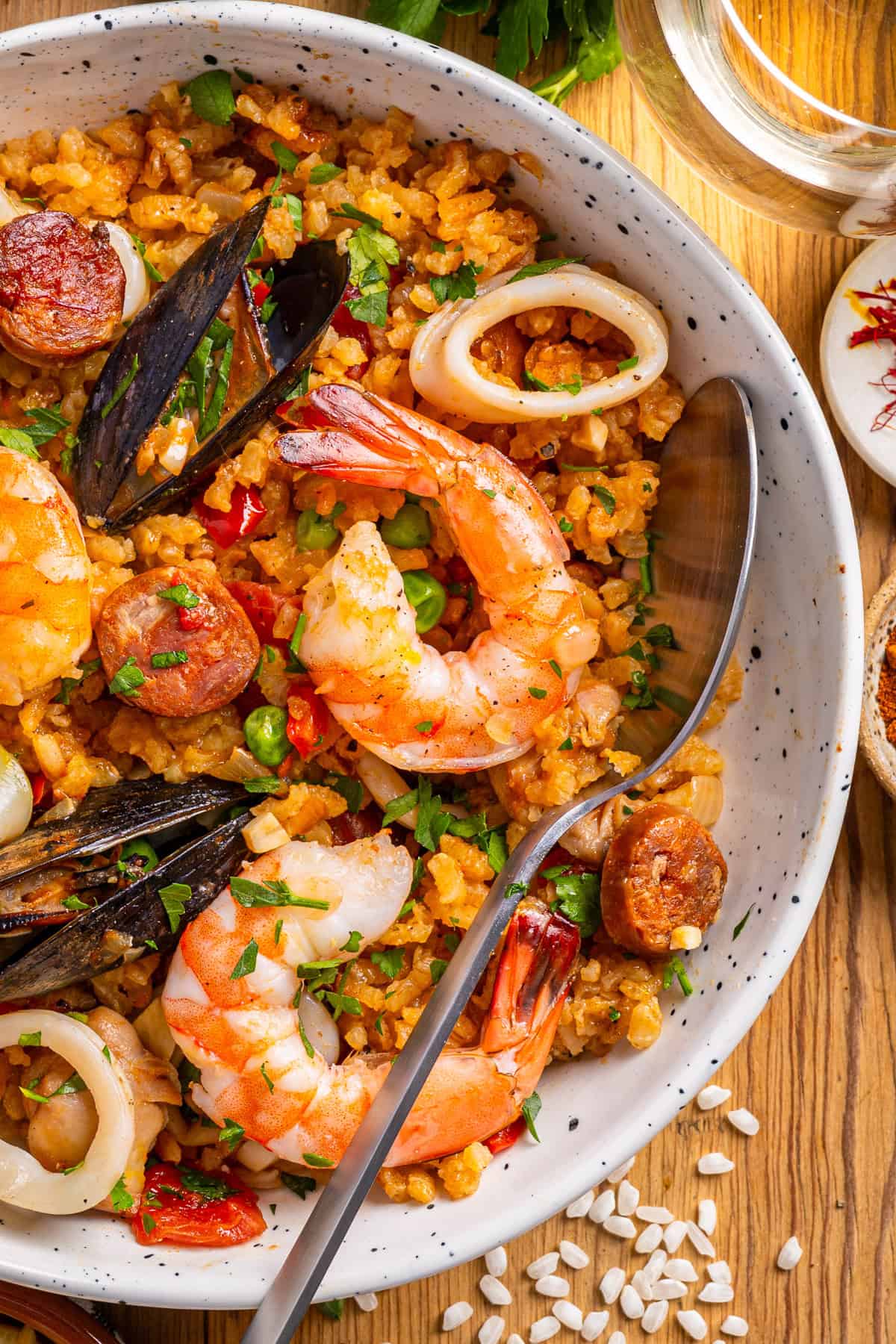 Paella A close-up of seafood paella with shrimp, mussels, squid rings, sliced sausage, peas, and rice in a speckled bowl, garnished with herbs.