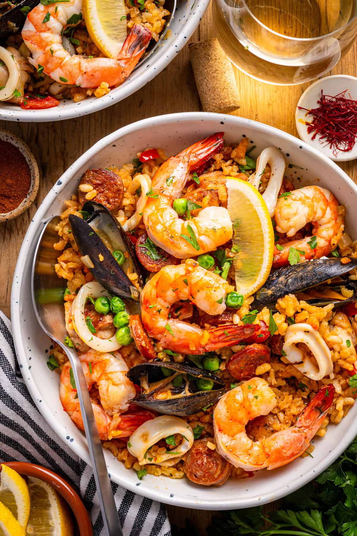 Paella A close-up of a bowl of seafood paella with shrimp, mussels, squid, chorizo, rice, peas, and lemon wedges.