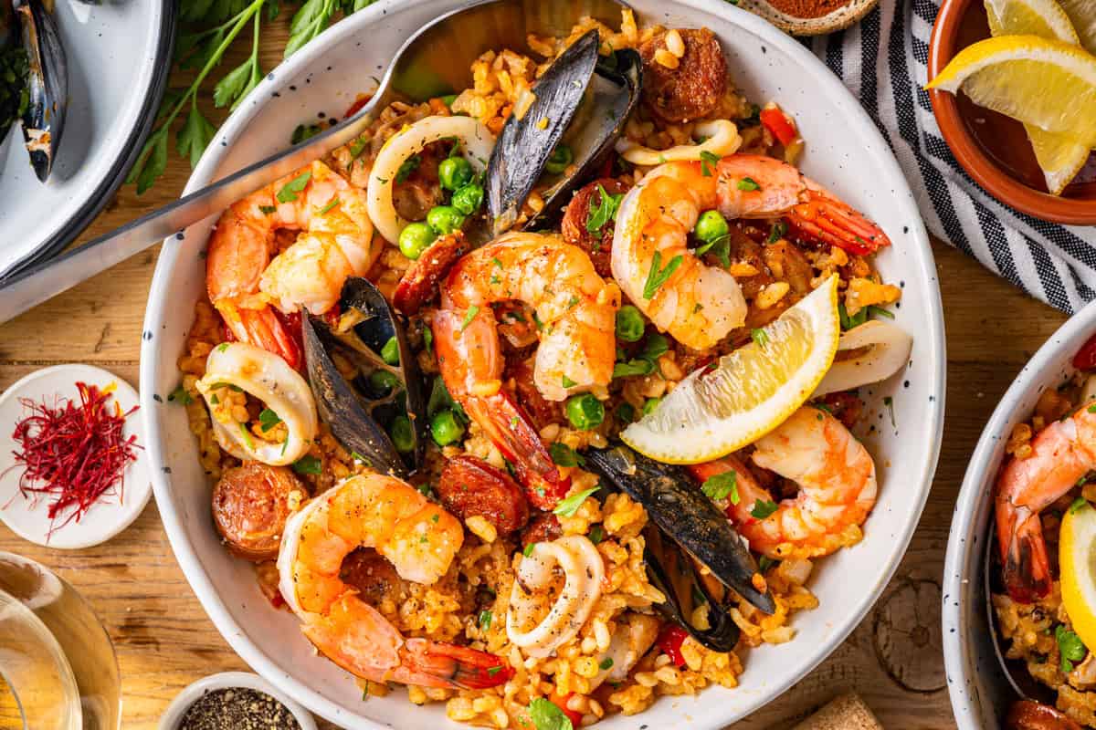 Paella A bowl of seafood paella with shrimp, mussels, squid, peas, rice, and chorizo, garnished with parsley and a lemon wedge.