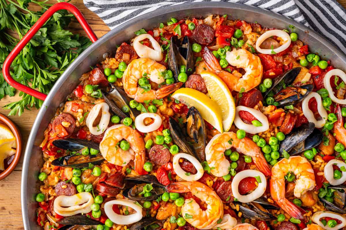 Paella A large pan of seafood paella topped with shrimp, mussels, calamari rings, chorizo, peas, and lemon wedges.