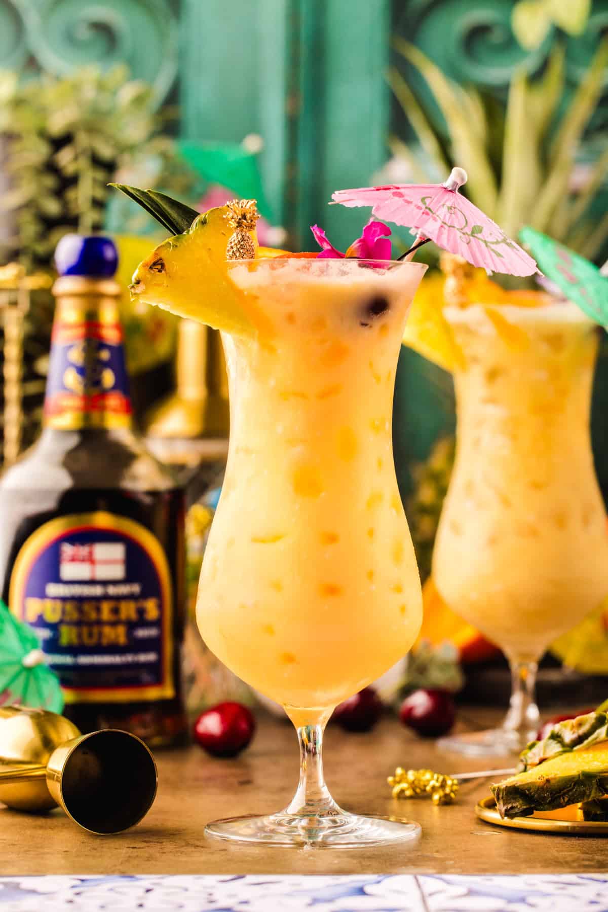Painkiller Cocktail A tropical yellow cocktail in a curvy glass with ice, pineapple slice, cherry, and a pink paper umbrella.