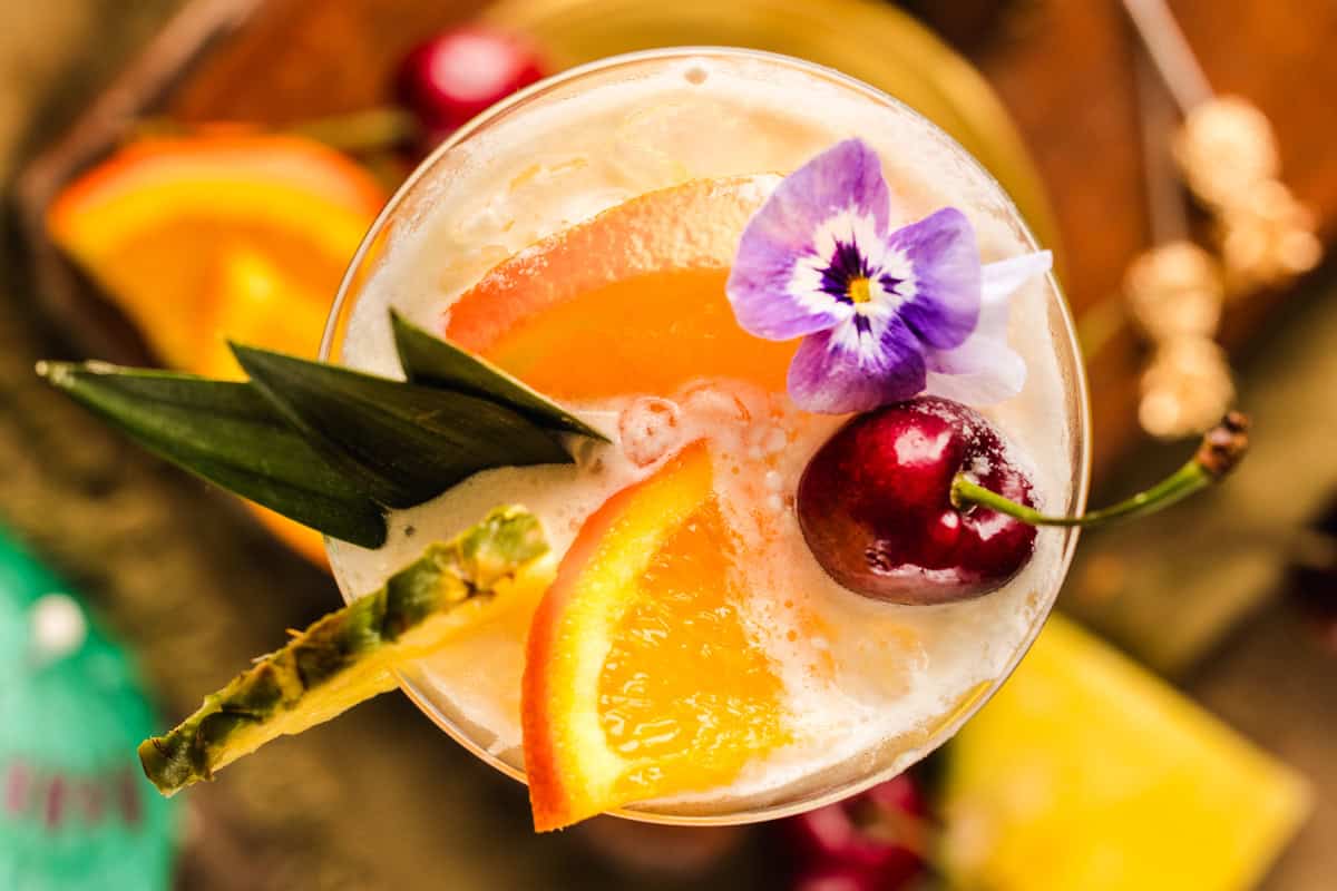 Painkiller Cocktail A tropical cocktail garnished with an orange slice, pineapple, a cherry, and a purple edible flower, viewed from above.