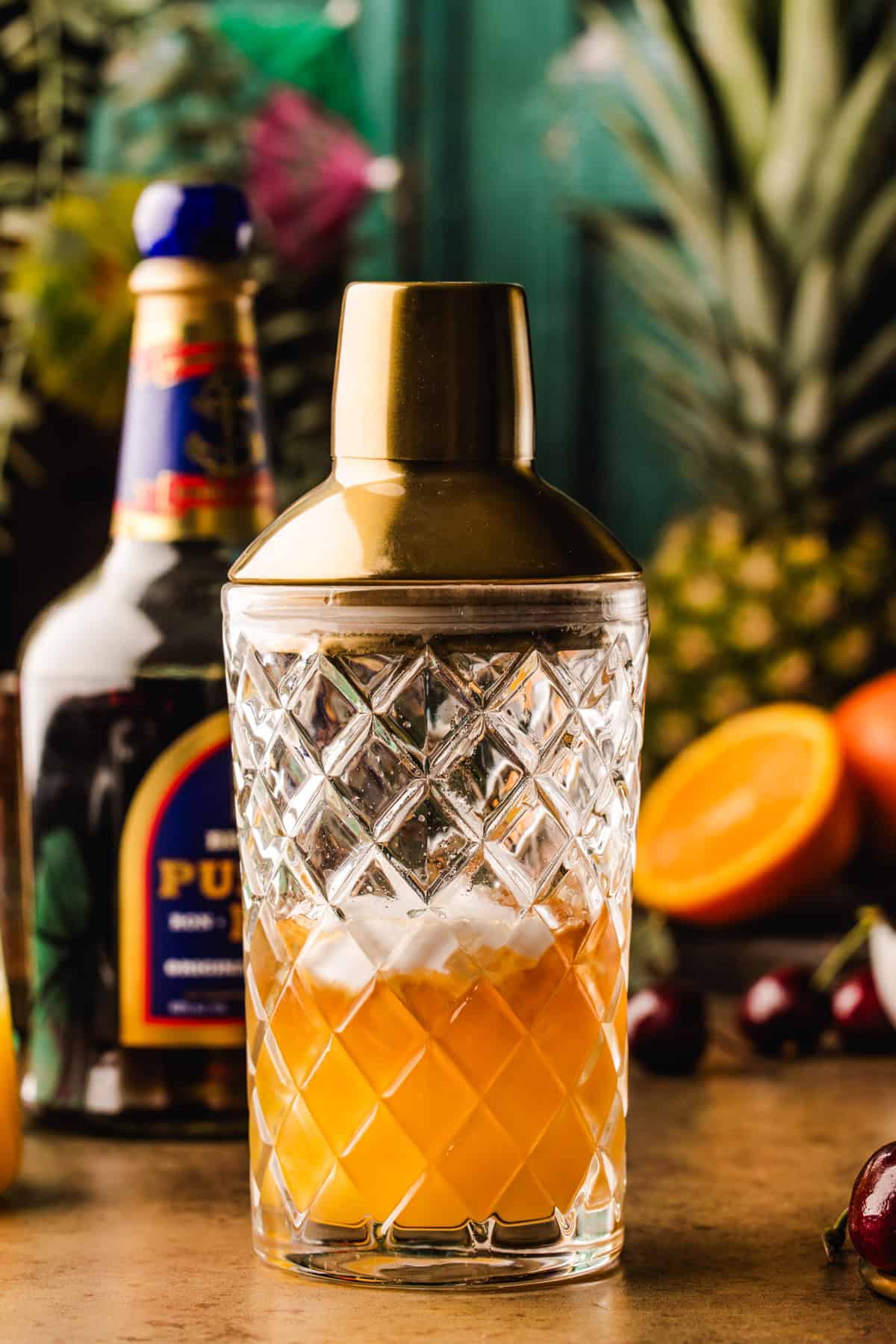 Painkiller Cocktail A crystal cocktail shaker with a gold lid filled with an yellowish drink sits on a table.