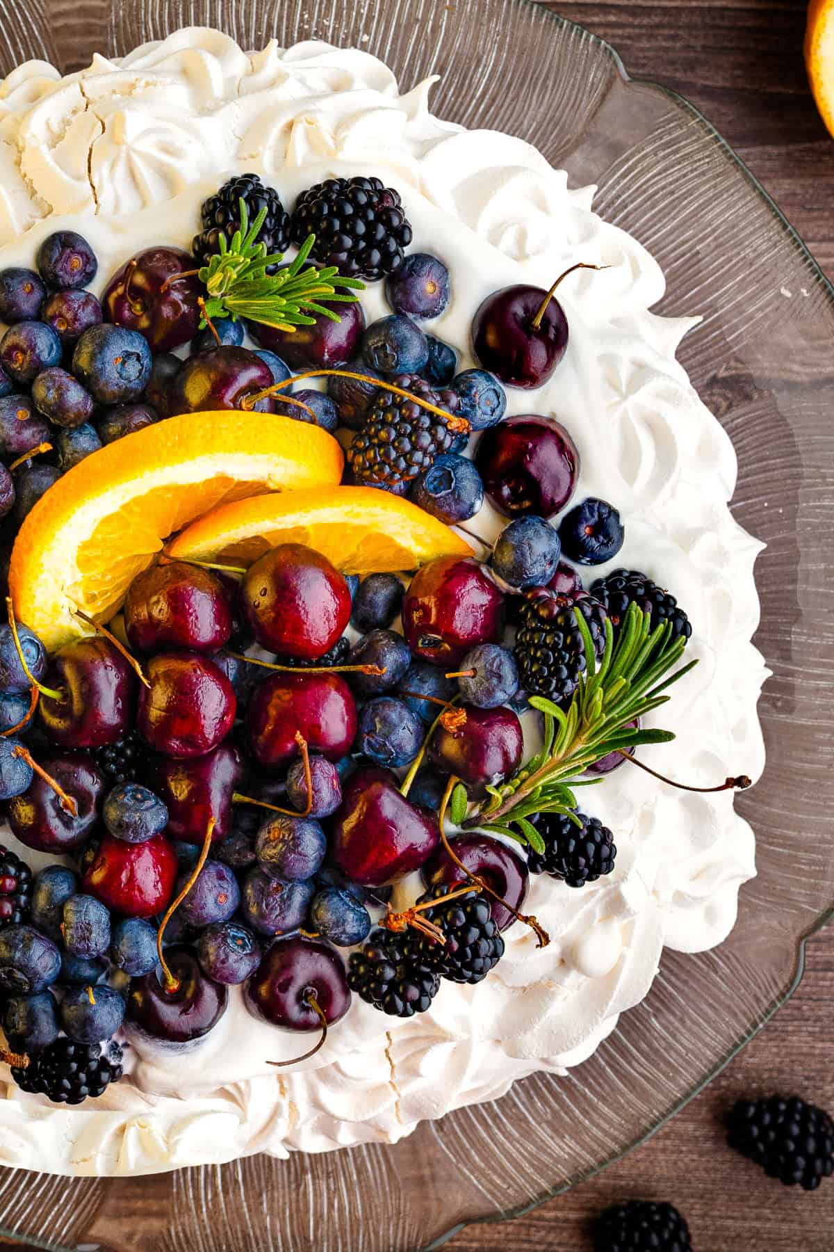 A pavlova topped with cherries, blueberries, blackberries, orange slices, and greenery, on a glass plate over a wooden table.