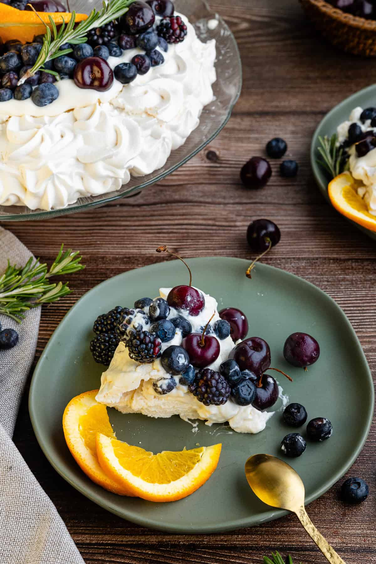 A slice of pavlova topped with cherries, blackberries, and blueberries sits on a green plate with orange slices. 