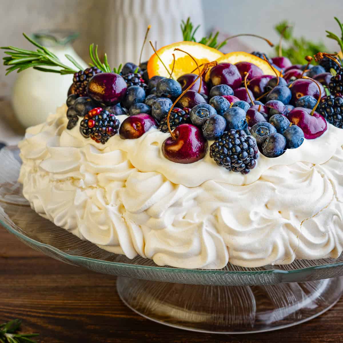 A pavlova dessert topped with fresh cherries, blueberries, blackberries, orange slices, and sprigs of rosemary sits on a glass cake stand. 