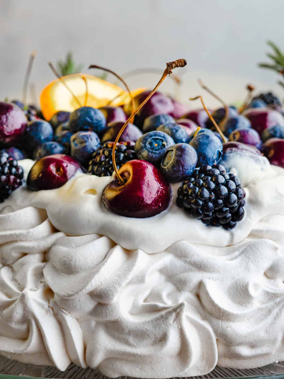 A pavlova dessert topped with whipped cream, cherries, blueberries, blackberries, and a slice of orange, with a swirled meringue base.