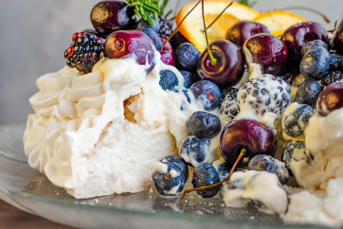 A close-up of a pavlova topped with whipped cream, blueberries, blackberries, cherries, and orange slices on a glass plate. 