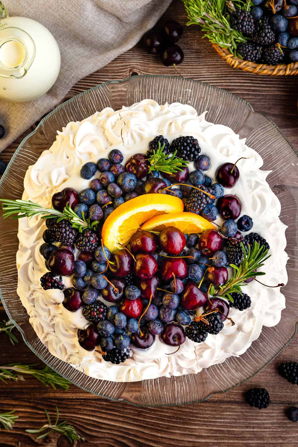 A pavlova topped with whipped cream, blueberries, blackberries, cherries, orange slices, and sprigs of rosemary.