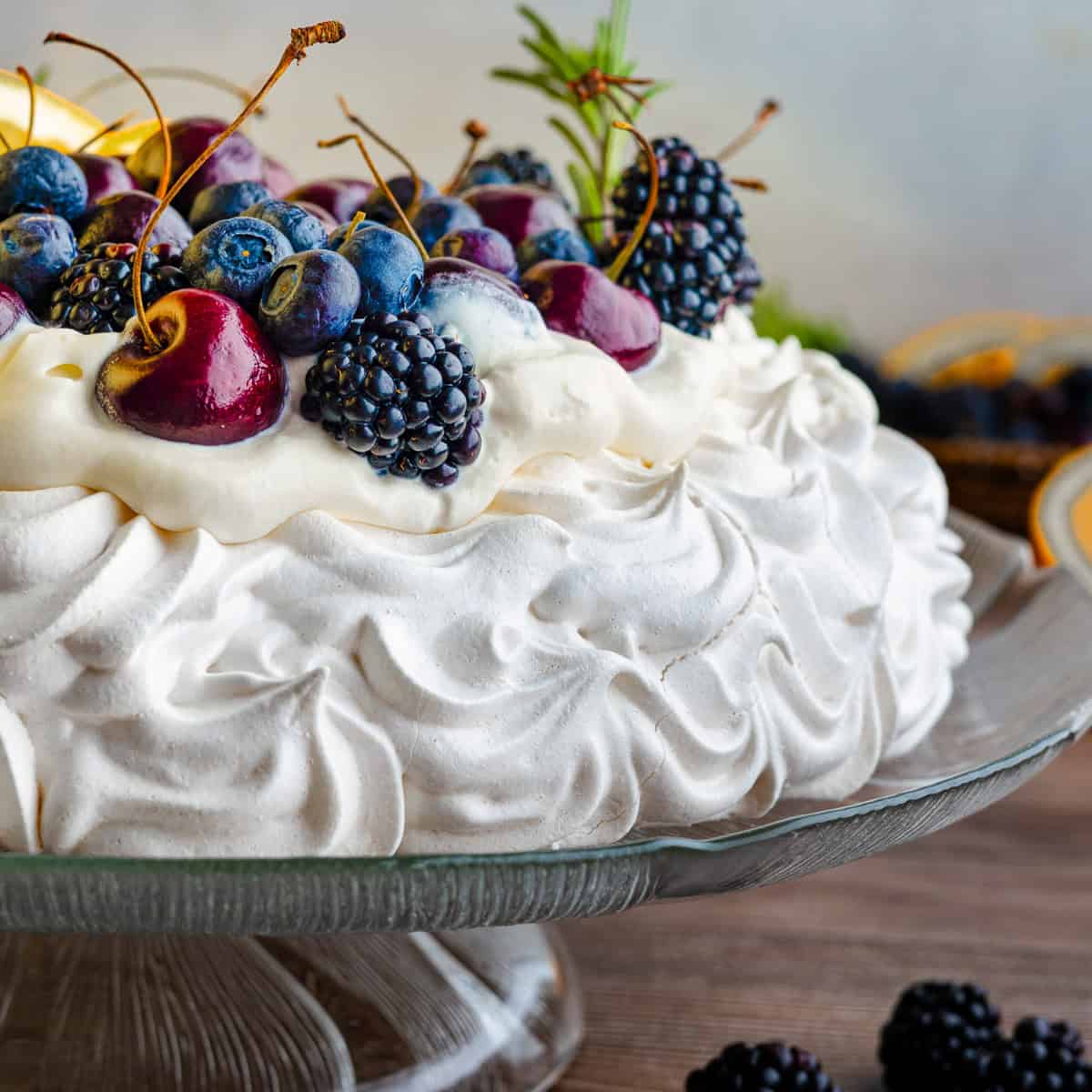 A pavlova topped with whipped cream, blueberries, blackberries, and cherries sits on a glass cake stand.