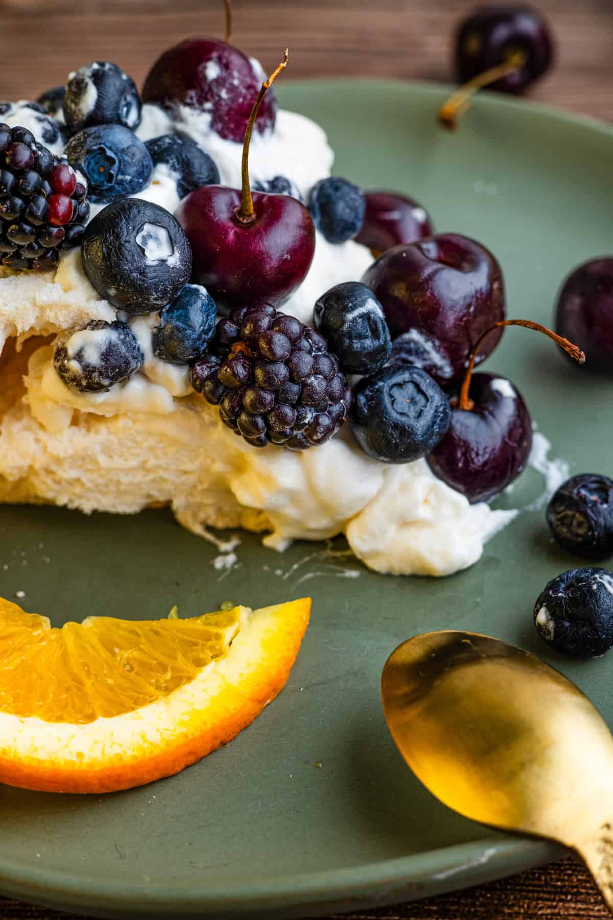 A close-up of a slice of Pavlova dessert topped with blueberries, blackberries, and cherries sits on a green plate with a gold spoon and an orange slice.