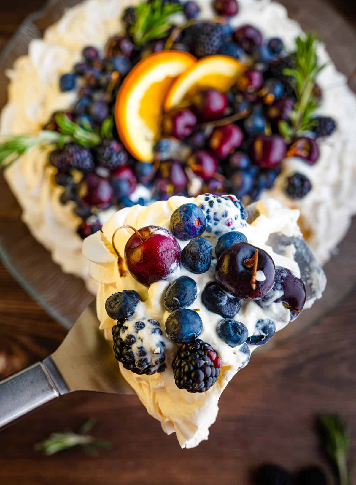 A slice of pavlova topped with whipped cream, cherries, blueberries, and blackberries is lifted from a whole pavlova garnished with fruit and orange slices.