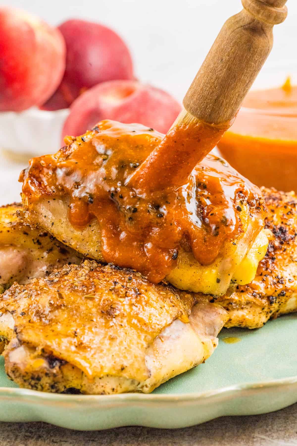 A wooden brush spreads thick, peach sauce over seasoned, cooked chicken thighs on a plate.