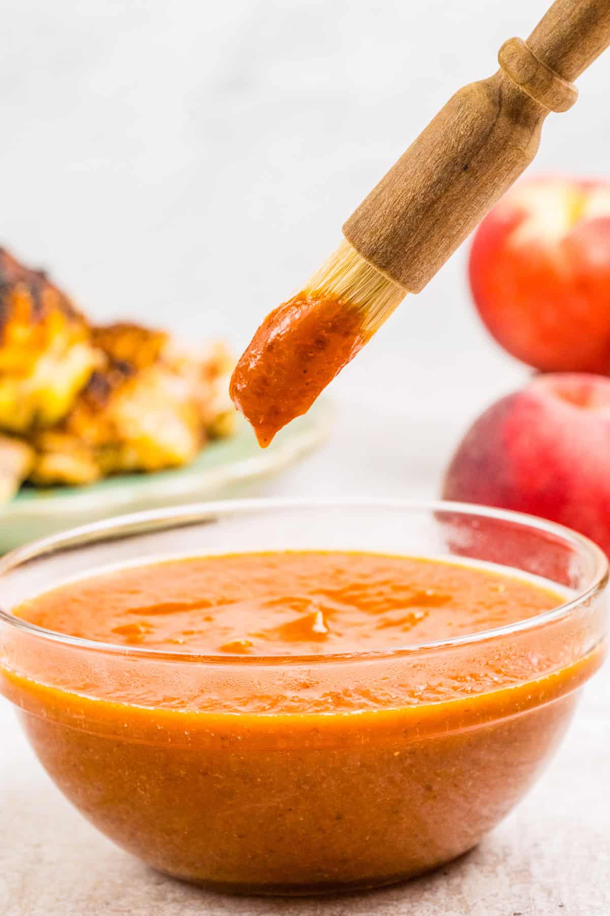 A bowl of peach sauce sits on a table. A basting brush coated in the sauce is held above the bowl. 