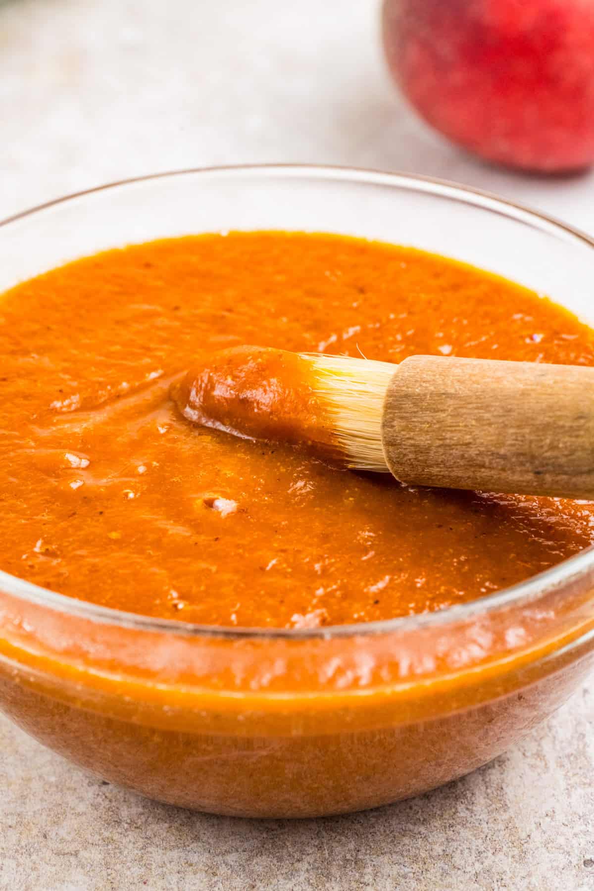 A glass bowl filled with peach sauce, with a basting brush resting on the rim and coated in the sauce. 