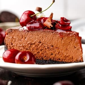 A slice of rich chocolate cheesecake with a dark crust, topped with fresh cherries, cherry sauce, and chocolate shavings.