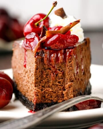 A slice of chocolate cheesecake topped with cherries, cherry sauce, whipped cream, and chocolate, served on a white plate with fresh cherries on the side.