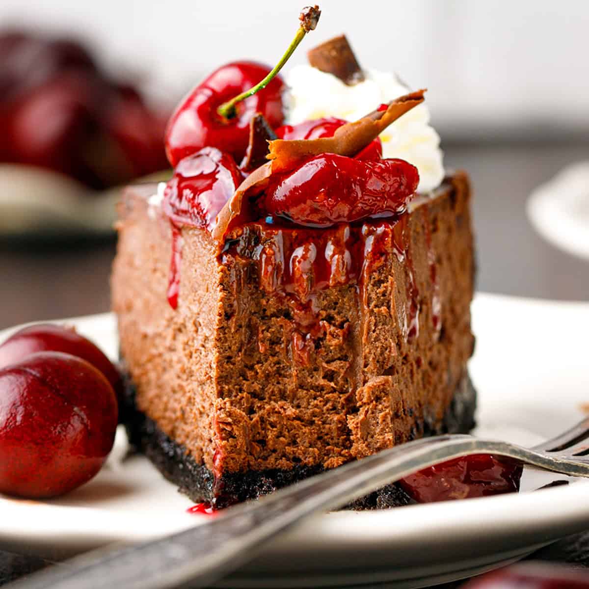A slice of chocolate cheesecake topped with cherries, cherry sauce, whipped cream, and chocolate, served on a white plate with fresh cherries on the side.