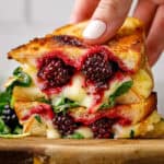 A hand holds a grilled cheese sandwich filled with melted cheese, blackberries, and greens, stacked on a wooden board.