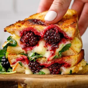 A hand holds a grilled cheese sandwich filled with melted cheese, blackberries, and greens, stacked on a wooden board.