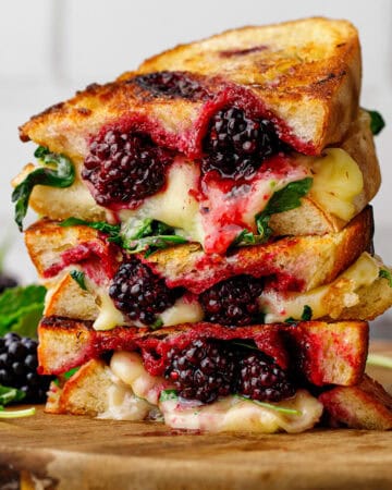 A stack of grilled cheese sandwiches filled with melted cheese, blackberries, and greens sits on a wooden board