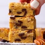 A hand holds the top piece of a stack of three blondie bars filled with chocolate chips and nuts.