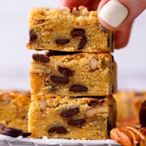A hand holds the top piece of a stack of three blondie bars filled with chocolate chips and nuts.