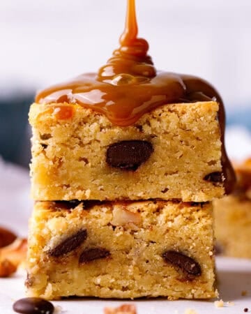 Two blondie dessert bars with chocolate chips are stacked. Thick caramel sauce is being poured over the top bar.