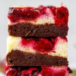 A close-up of three stacked brownies with creamy cheesecake and vibrant red berry swirl layers, showing a rich, moist texture.
