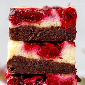 A close-up of three stacked brownies with creamy cheesecake and vibrant red berry swirl layers, showing a rich, moist texture.