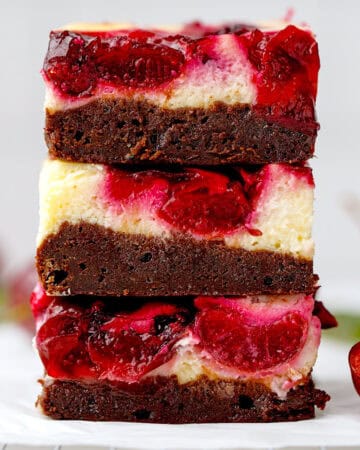 Three stacked dessert bars with a bottom layer of rich chocolate brownie, a creamy cheesecake layer, and a topping of glossy red cherries.
