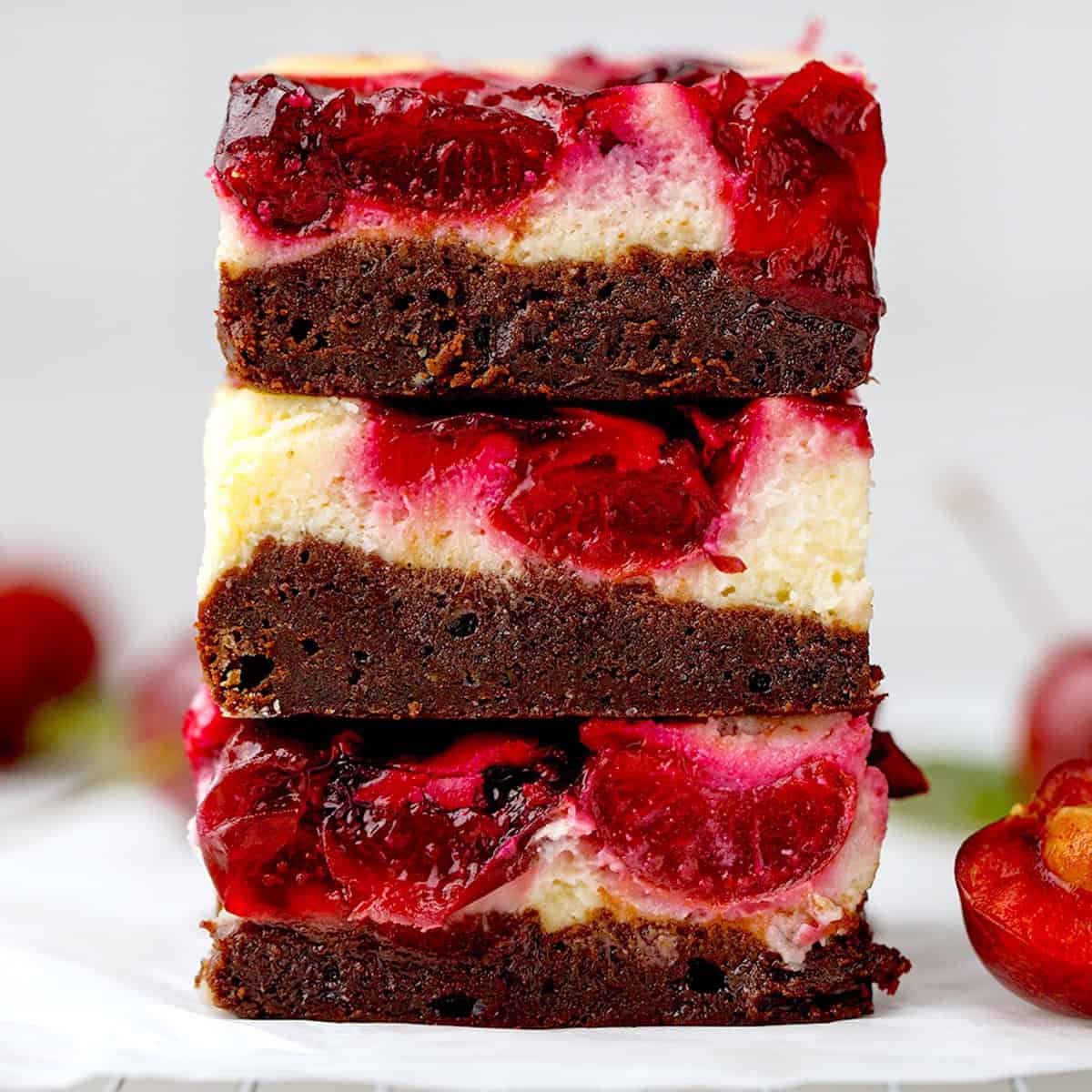 Three stacked dessert bars with a bottom layer of rich chocolate brownie, a creamy cheesecake layer, and a topping of glossy red cherries.