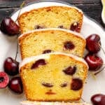 Three slices of glazed cherry loaf cake are arranged on a white plate, surrounded by fresh cherries.