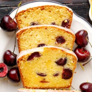 Three slices of glazed cherry loaf cake are arranged on a white plate, surrounded by fresh cherries.