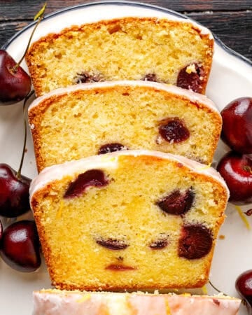 Three slices of glazed cherry loaf cake are arranged on a plate, surrounded by fresh cherries.