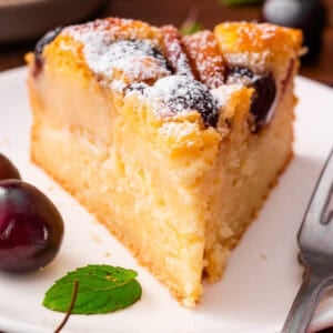 A slice of cherry cake dusted with powdered sugar on a white plate, with fresh cherries and a mint leaf beside it, and a fork nearby.