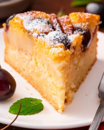 A slice of cherry cake dusted with powdered sugar on a white plate, with fresh cherries and a mint leaf beside it, and a fork on the side.