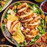 A salad with sliced grilled chicken, arugula, pear slices, walnuts, dried cranberries, and crumbled cheese, served in a bowl with a fork.