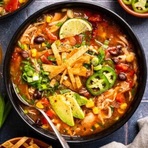 A bowl of chicken tortilla soup topped with avocado slices, jalapeños, tortilla strips, lime, green onions, and cilantro, with colorful vegetables and black beans visible in the broth.