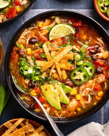 A bowl of chicken tortilla soup topped with avocado slices, crispy tortilla strips, jalape&ntilde;o, lime, green onions, corn, black beans, and tomatoes, with a spoon and fresh ingredients on the side.