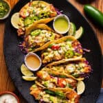 Fish tacos topped with sliced jalapeños, mango salsa, cheese, and avocados are arranged on a black platter, garnished with lime wedges.