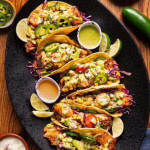 Fish tacos topped with sliced jalape&ntilde;os, mango salsa, cheese, and avocados are arranged on a black platter, garnished with lime wedges.