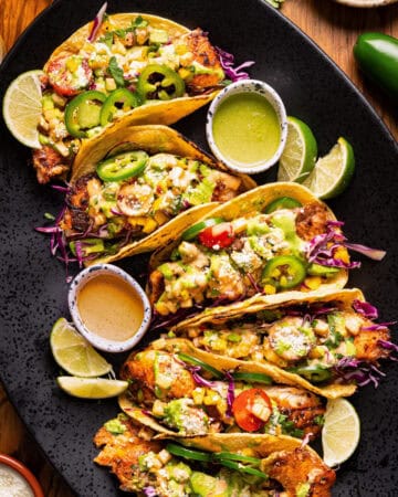 Crispy tacos filled with vegetables and topped with salsa, cheese, and jalape&ntilde;os, are arranged on a black platter with lime wedges and sauces on the side.