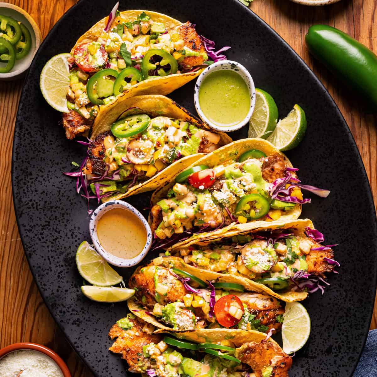 Crispy tacos filled with vegetables and topped with salsa, cheese, and jalapeños, are arranged on a black platter with lime wedges and sauces on the side.