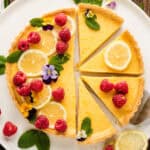 A lemon tart with slices cut out, decorated with lemon slices, raspberries, edible flowers, and mint leaves on a white plate.
