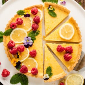 A lemon tart with slices cut out, decorated with lemon slices, raspberries, edible flowers, and mint leaves on a white plate.